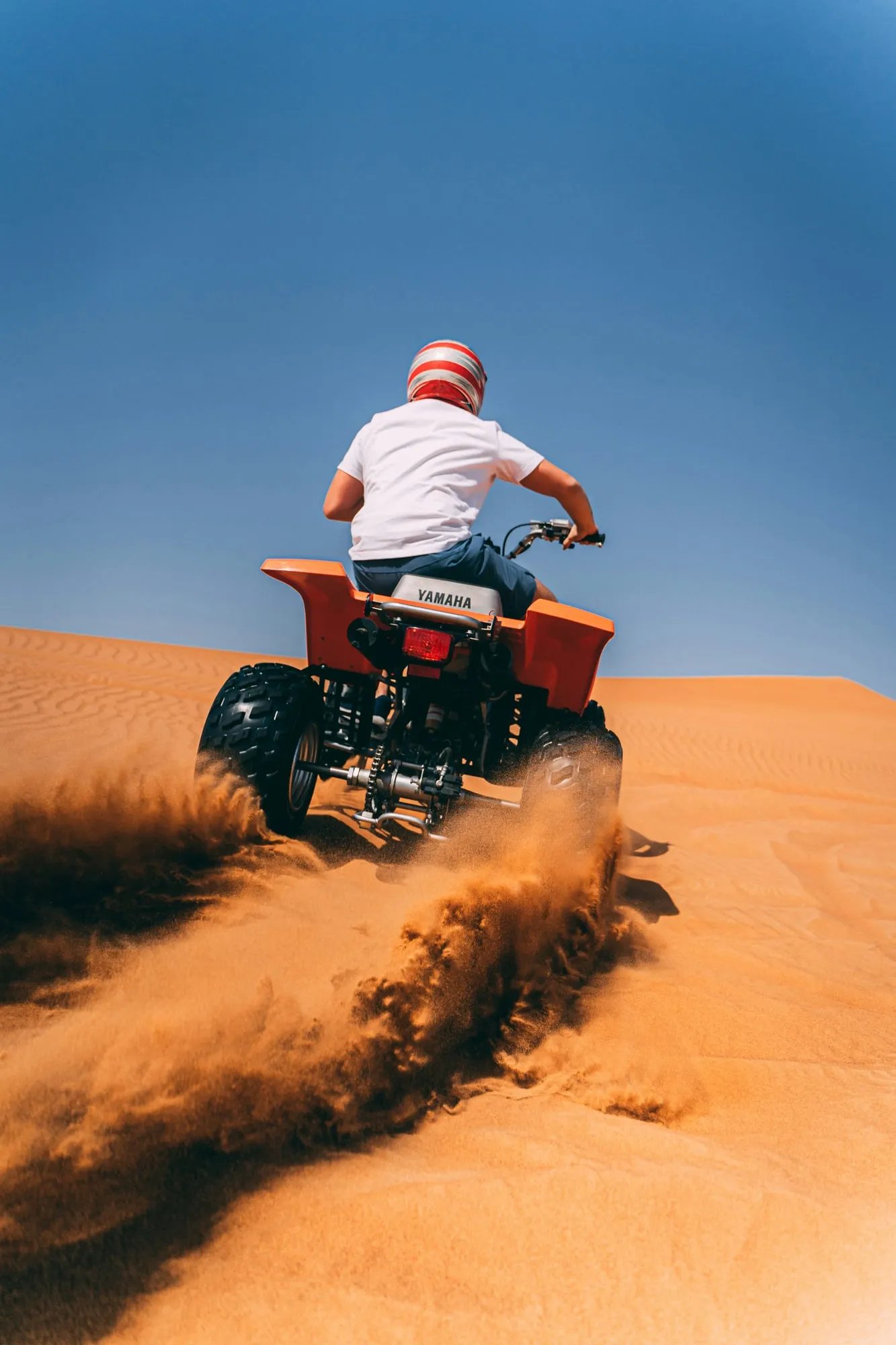 Riding a quad bike up a dune, sand flying, under a clear blue sky